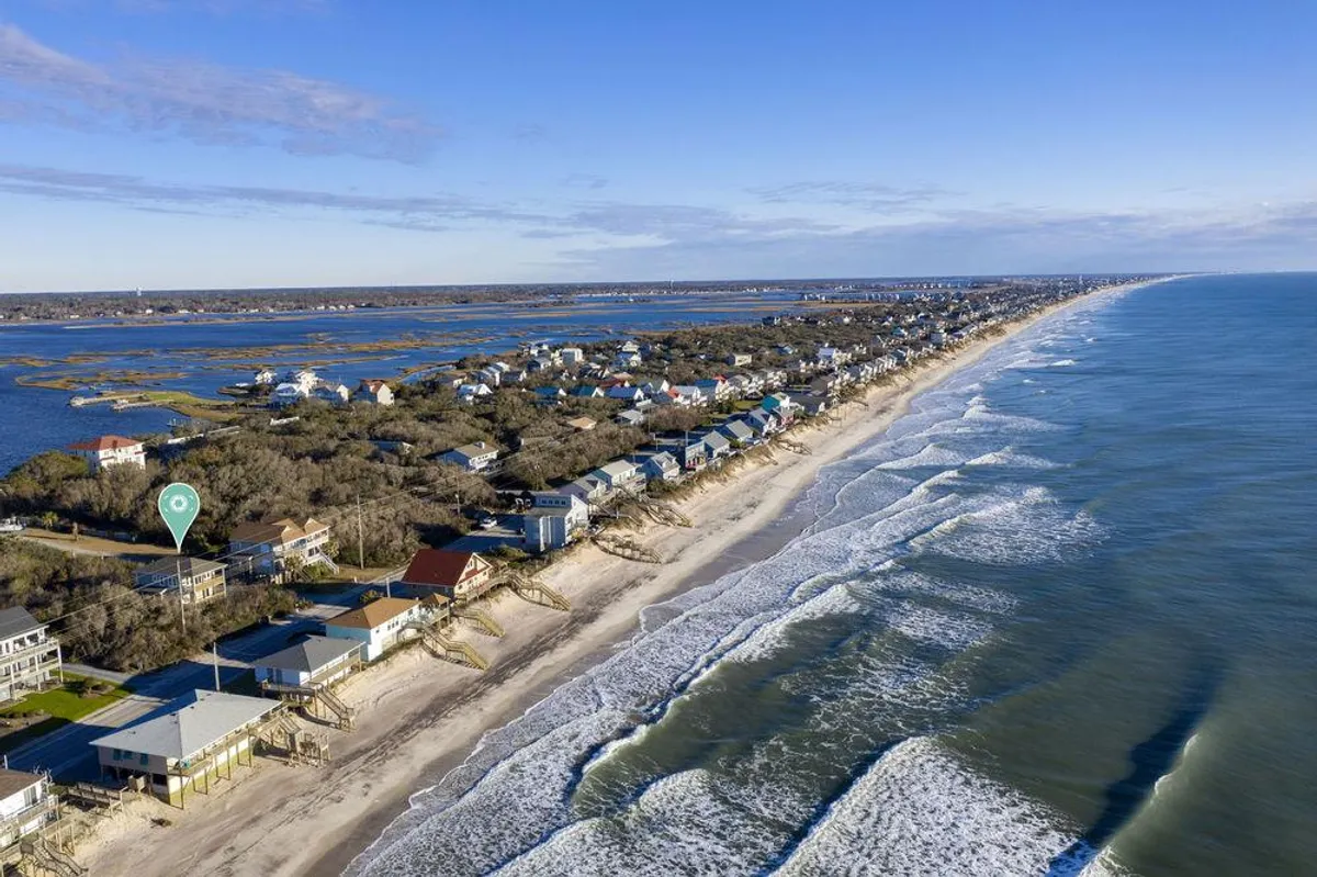An elevated perspective of the coastline, featuring ocean waves, beachfront houses, lush marshlands, and the distant horizon. The image captures both the wild natural aspects and the inviting residential environment.