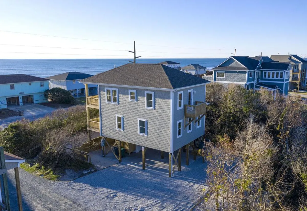 The image showcases a charming single-family beach house with a spacious deck, just steps from the shoreline. The ocean is visible in the background, and the deck overlooks both the waves and the tranquil sound. The scene evokes a peaceful, inviting atmosphere perfect for relaxing, watching sunsets, or gathering with family and friends.