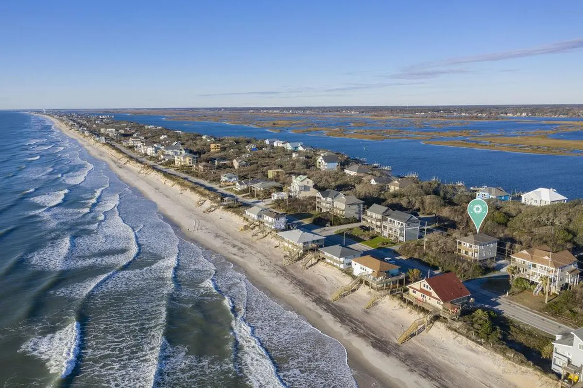A wide view of the expansive sandy beach, shimmering ocean waves, coastal homes lining the shore, and a calm waterway in the distance. The scene is vibrant with natural beauty and hints at the many water-based and outdoor activities available in the area.
