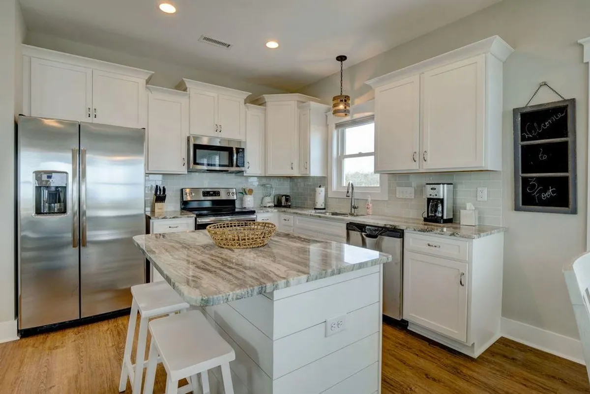 This image features a welcoming kitchen centered around an island countertop, complete with stainless steel appliances, a stylish hanging light fixture, and a chalkboard sign. The open layout is designed for cooking, casual breakfasts, and socializing, allowing for seamless interaction among guests.