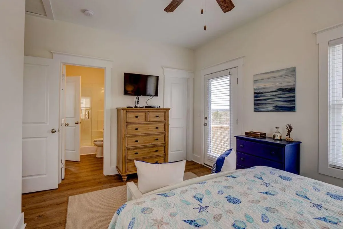 A tranquil bedroom featuring a bed with a patterned quilt, a wooden dresser, a blue accent dresser, a wall-mounted TV, and an open door leading out to a balcony. Sunlight streams through the doorway, inviting the outdoors in and providing an airy, restful atmosphere.