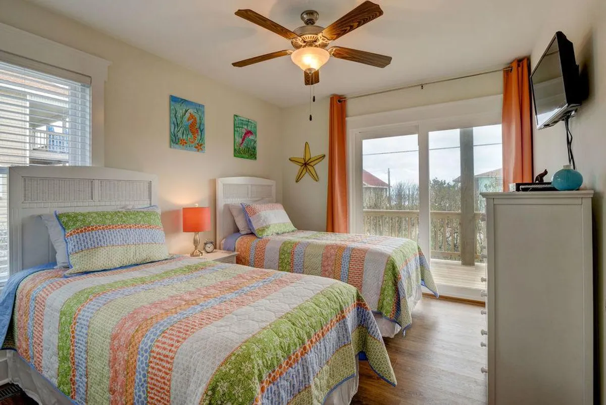 A cheerful twin bedroom with two beds dressed in colorful quilts, bright wall art, a ceiling fan, and large windows that open directly to a balcony. The space radiates playful relaxation, ideal for children or guests seeking a cozy retreat and fresh air.