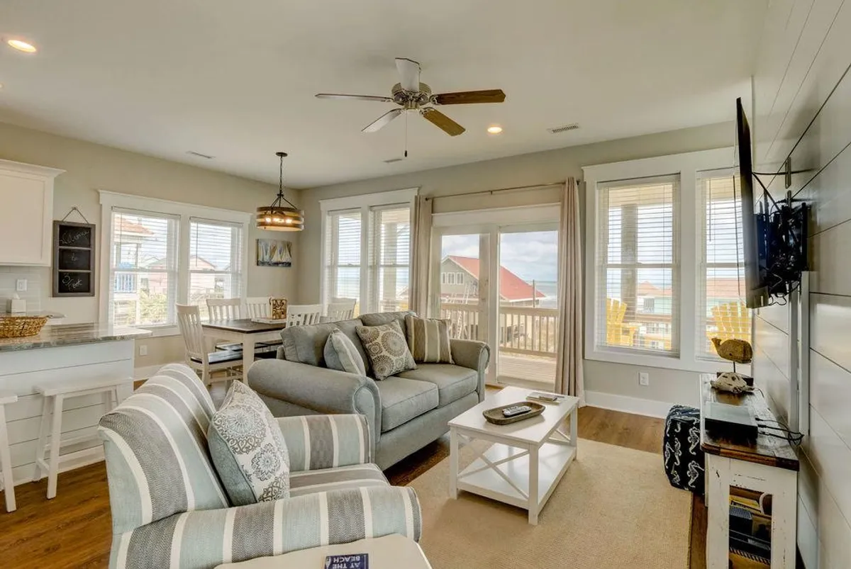 A cozy living space featuring a plush sofa adorned with decorative pillows, a dining table set nearby, large windows with soft curtains, and a wall-mounted TV. Natural light fills the room, creating an inviting atmosphere for relaxation and entertainment.