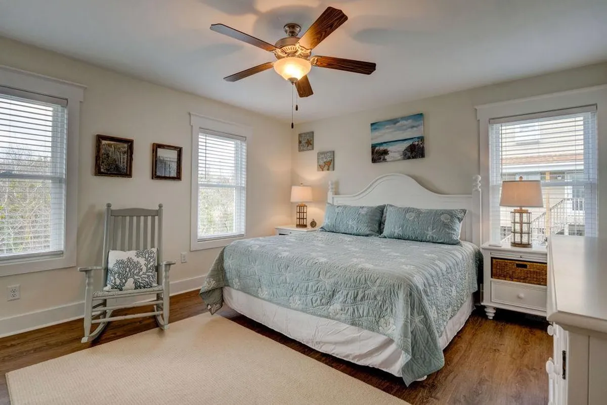 A serene bedroom featuring a bed with a decorative quilt, a rocking chair next to a nightstand and lamp, and charming wall art. The space is bathed in natural light, evoking a sense of calm and restful privacy.
