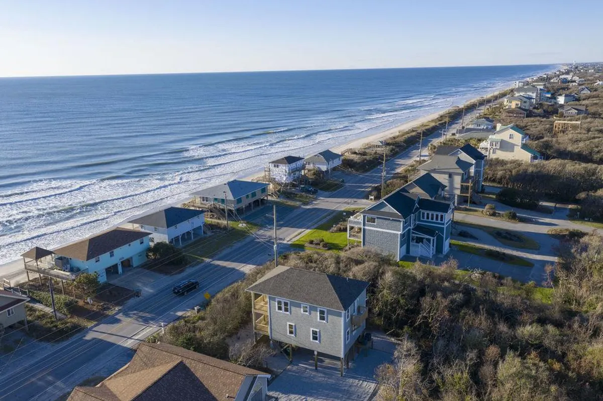 A sweeping exterior view shows gentle ocean waves rolling onto the sandy shoreline, with rows of beachfront houses and a winding roadway framed by lush coastal vegetation. The scene captures the vibrant outdoor life, inviting families to beach walks, fishing, or soaking in the coastal atmosphere.