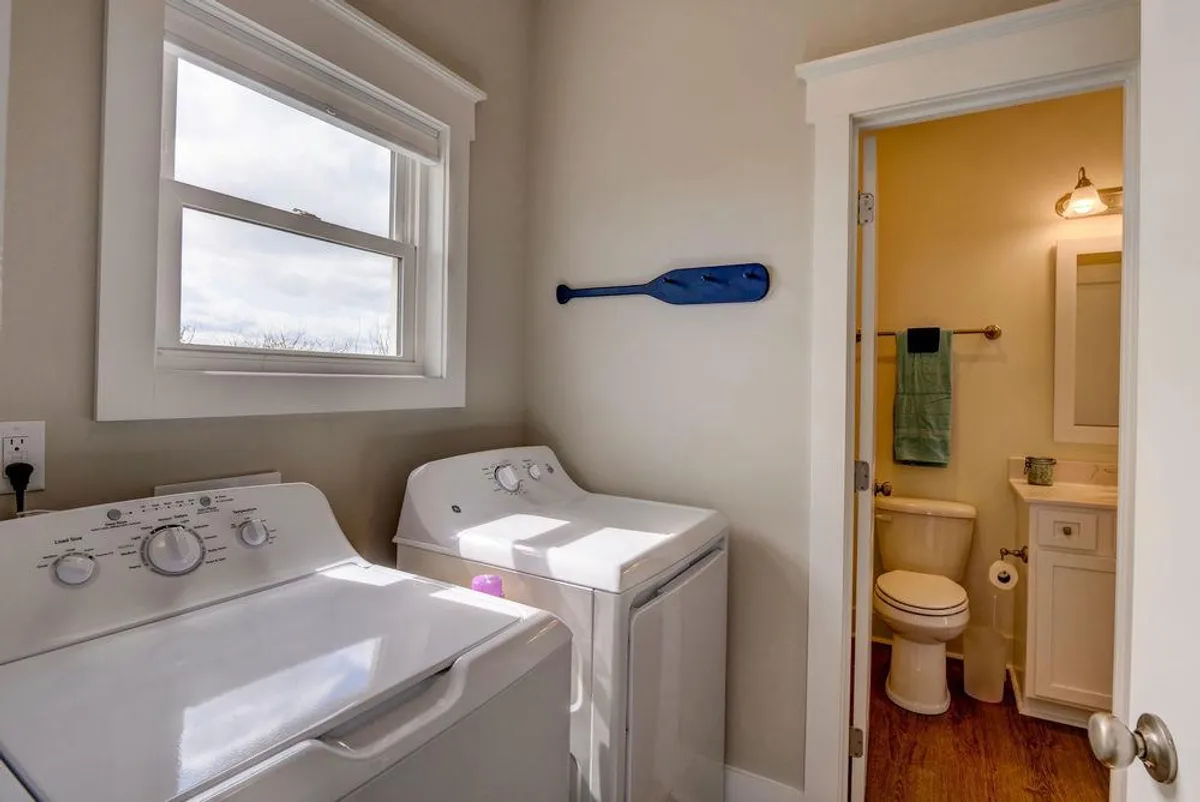 A functional and organized laundry area with a washing machine, dryer, and a bathroom entrance nearby. Decor includes a blue paddle, adding a coastal touch to the practical setup.